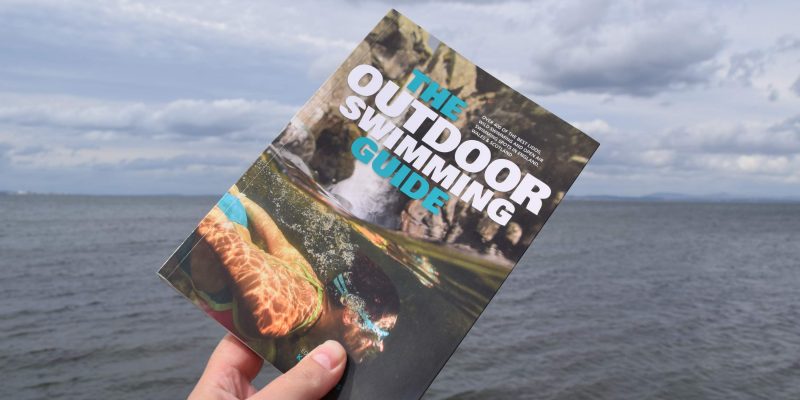Hand holding The Outdoor Swimming Guide in front of the sea